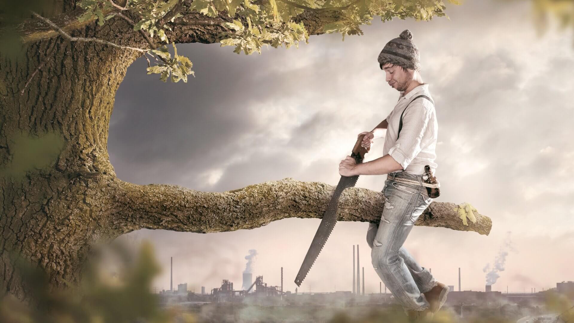 A man sits on the wrong side of a tree branch, holding a saw.