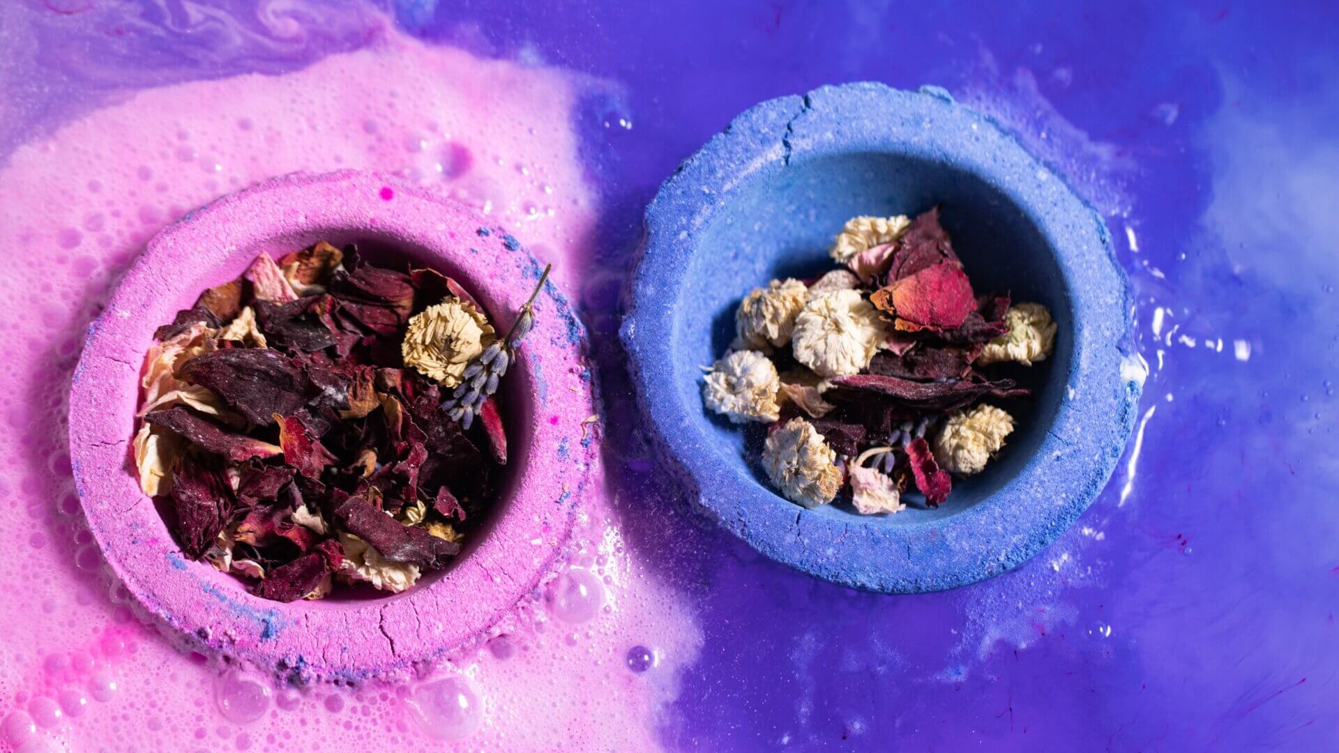 Two bathbombs in water half and half one pink and one purple with flower petals in.